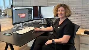 ד"ר שרית בן שמן, אורתודונטית, יושבת ליד שולחן משרדי עם שני מסכים. על המסך השמאלי מוצגת סריקה דיגיטלית תלת-ממדית של לסת שיניים לפני טיפול אורתודונטי. ד"ר בן שמן מחייכת ומחזיקה ביד משקפיים.