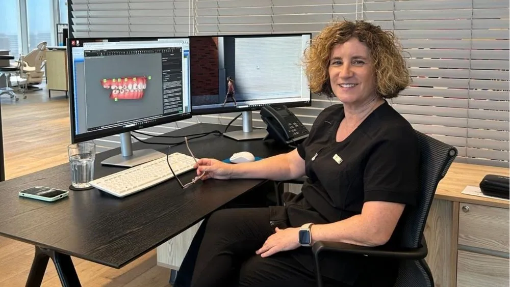 ד"ר שרית בן שמן, אורתודונטית, יושבת ליד שולחן משרדי עם שני מסכים. על המסך השמאלי מוצגת סריקה דיגיטלית תלת-ממדית של לסת שיניים לפני טיפול אורתודונטי. ד"ר בן שמן מחייכת ומחזיקה ביד משקפיים.