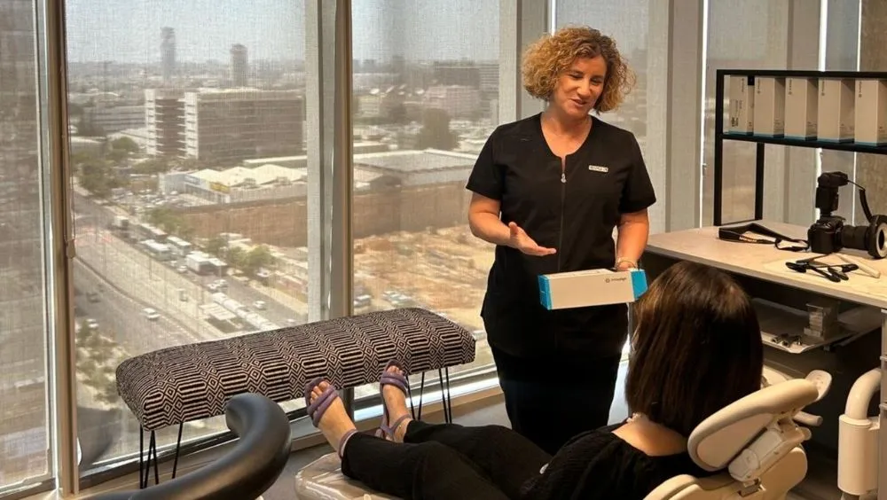 ד"ר שרית בן שמן, אורתודונטית, מחייכת ומציגה למטופלת ששוכבת על כיסא טיפולים את ערכת יישור השיניים השקופה של אינויזליין (Invisalign). ברקע רואים נוף אורבני של העיר דרך חלון גדול.